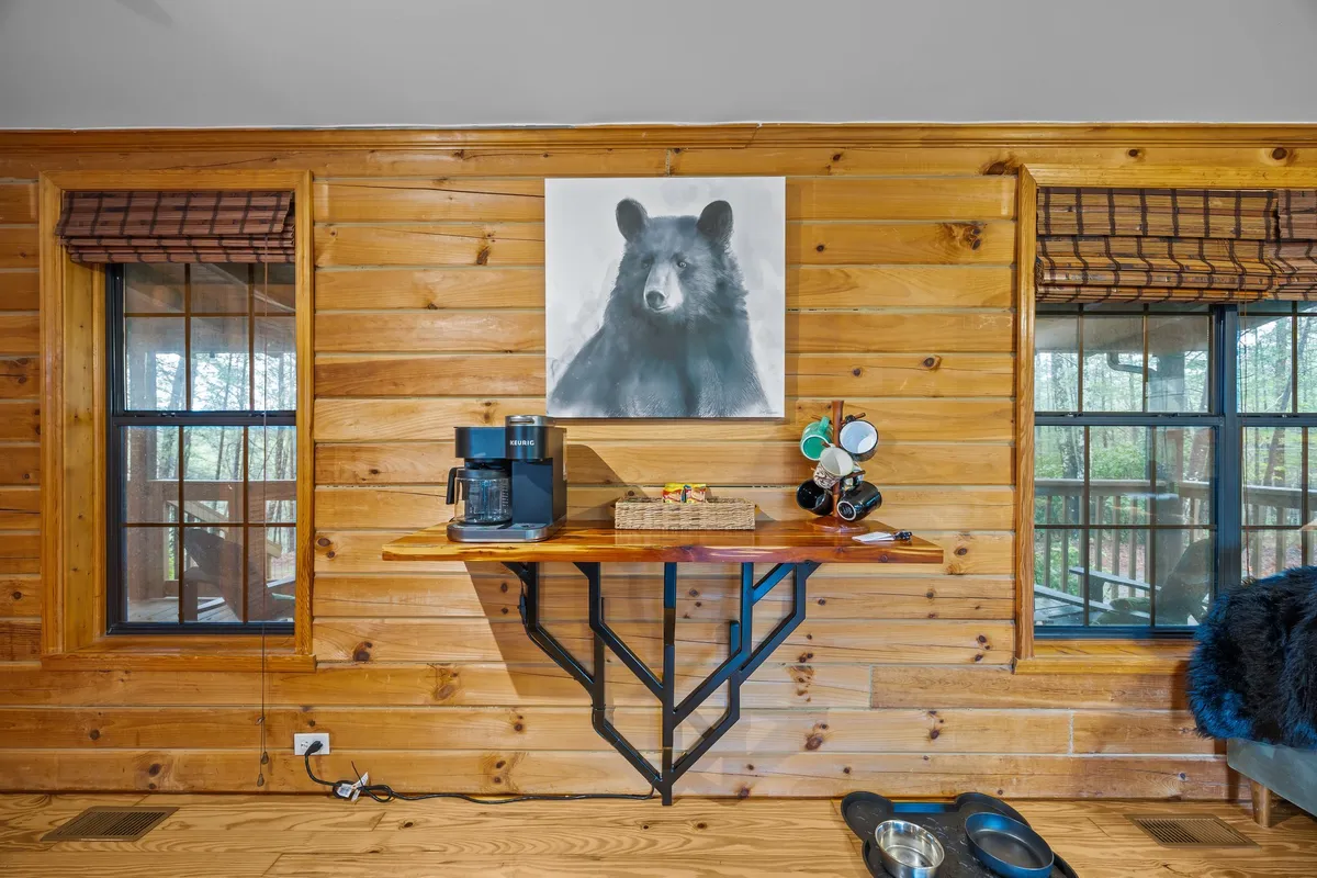 A cozy interior space with natural wooden walls, a charming bear painting, and a coffee station set near windows. Through the windows, guests can glimpse the surrounding greenery and potentially spot wildlife. The setup invites moments of relaxation while connecting with the outdoors.