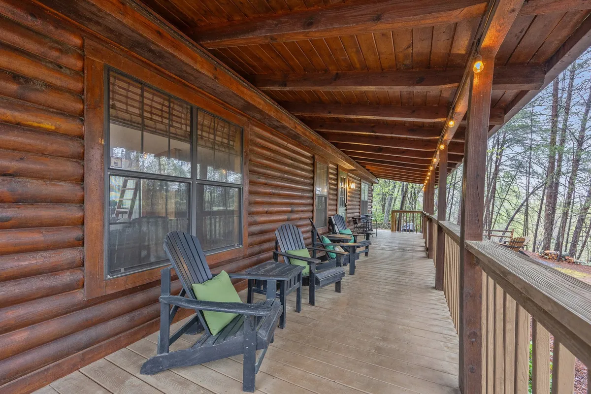 A rustic wooden porch features rocking chairs with green cushions, set against a log cabin exterior and surrounded by trees. The setting is perfect for unwinding with a book, sharing a glass of wine, or watching the sunset together, offering views of starlit skies by night.