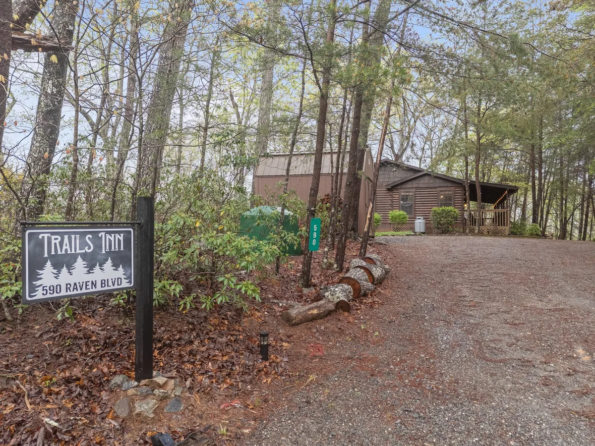 This inviting exterior shot features the Trails Inn sign at the property entrance, with a gravel driveway leading to a wooden porch surrounded by lush trees. The scene evokes a welcoming, rustic atmosphere and suggests a place ideal for gathering and beginning outdoor adventures.