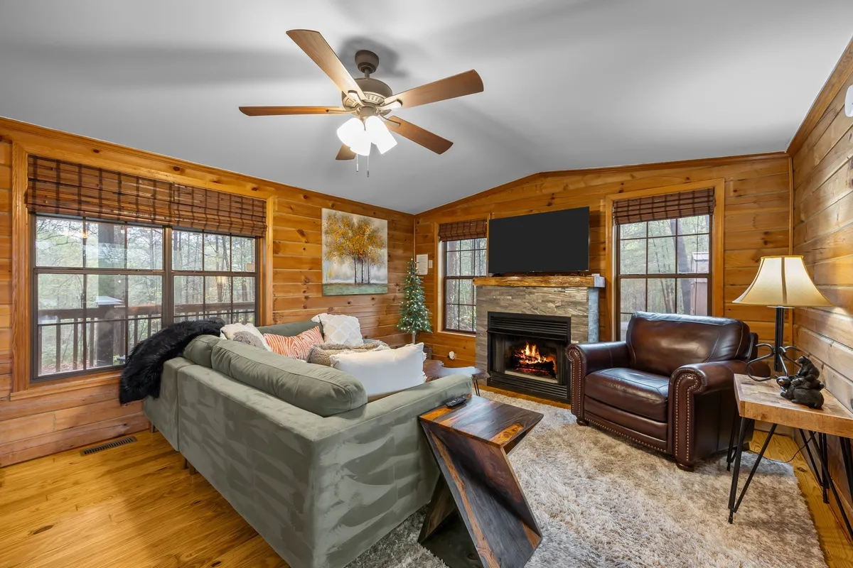 A welcoming living room centered on a striking stone fireplace, flanked by a comfortable sofa and artwork on the walls. Large windows let in natural light and frame serene outdoor views, while the decor subtly nods to the surrounding mountains with its earthy tones and cozy touches.