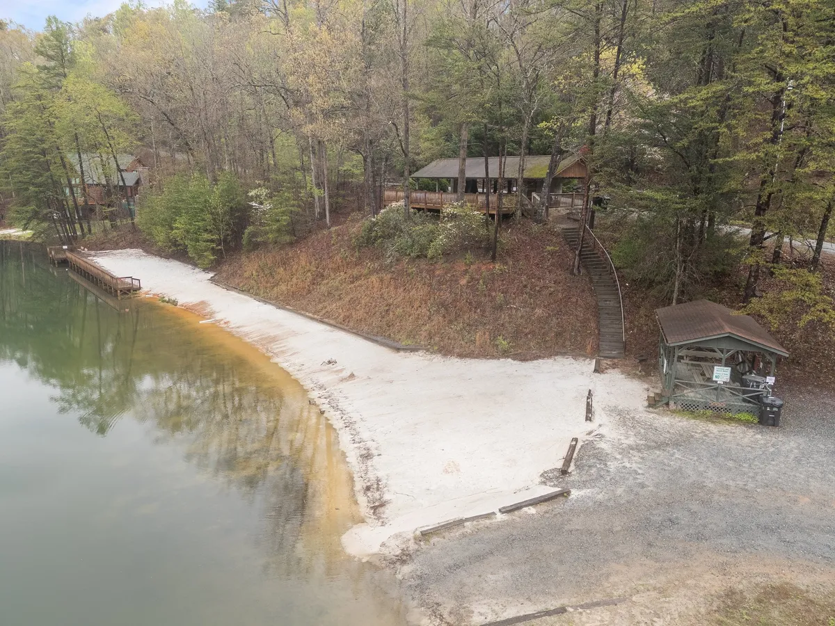 This scene captures a tranquil lake bordered by trees and a soft beach, with a wooden deck and shaded gazebo overlooking the water. The setting is ideal for lakeside activities like kayaking, fishing, or relaxing in a serene environment.