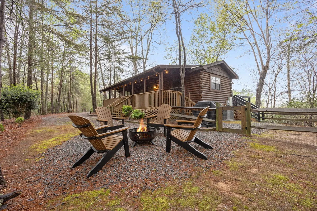 The hero image showcases the log cabin's inviting exterior, featuring a glowing fire pit surrounded by wooden chairs. The cozy setup suggests intimate gatherings, with the backdrop of lush forest and a hint of lake tranquility nearby. The scene evokes warmth, relaxation, and togetherness under a starlit sky.