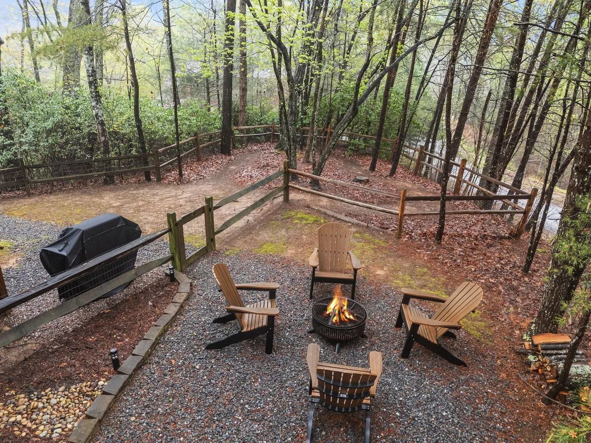 The photograph showcases an inviting fire pit encircled by Adirondack chairs, with a grill nearby and trees providing a natural backdrop. The scene evokes warmth and camaraderie, ideal for gathering with friends or family in the evening.