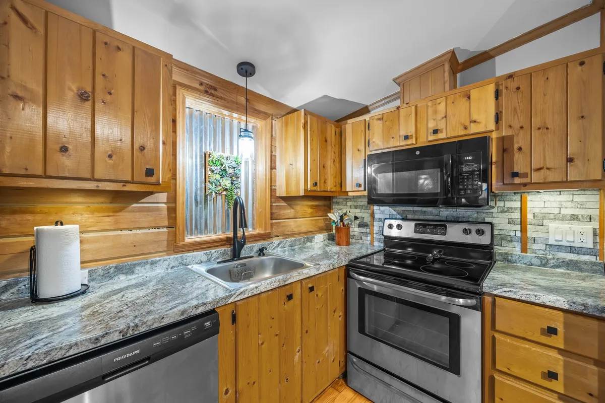 This image reveals a warmly lit kitchen featuring rustic wooden cabinets, sleek stainless steel appliances, and a granite countertop. A window above the sink offers glimpses of surrounding greenery, enhancing the cozy, welcoming atmosphere. The space looks ready for preparing hearty meals or gathering for conversation around the counter.